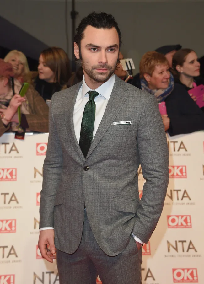 Man in a gray suit with a green tie at a red carpet event, surrounded by people taking photos.