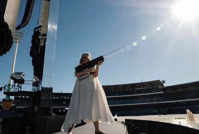 A person in a white dress stands on a stage holding a large tube, with a stadium and bright sun in the background.
