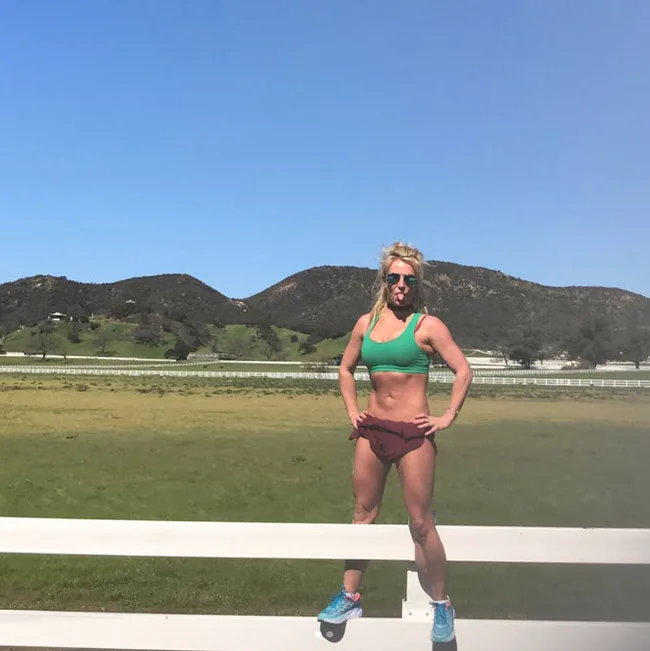 Person in workout clothes stands on a fence, tongue out, with a field and hills in the background on a sunny day.
