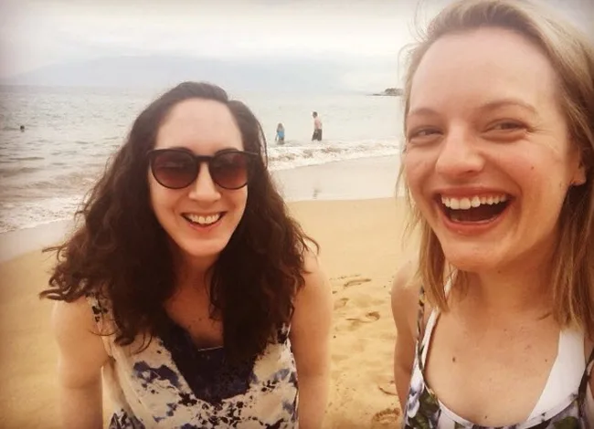 Two women smiling on a beach, one wearing sunglasses. Ocean waves and distant people are visible in the background.