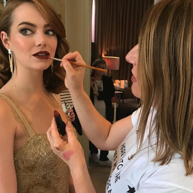 Woman getting makeup applied by a makeup artist, wearing a gold dress, with a focus on lipstick application.