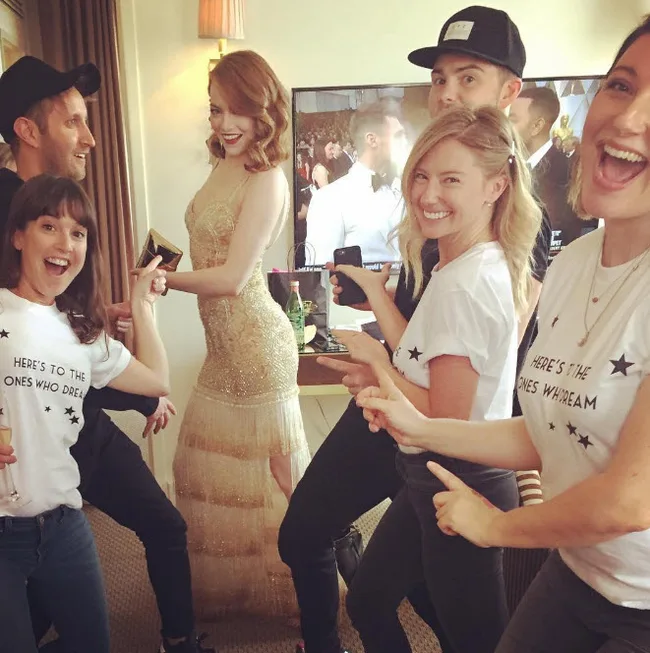 Group celebrating in a room with a woman in a gold dress, some wearing matching shirts that say "Here's to the ones who dream."