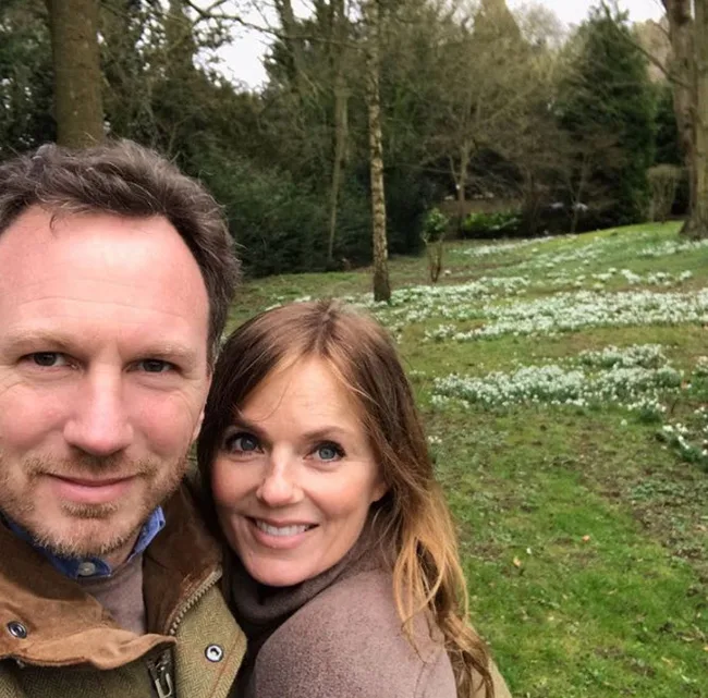 A couple smiling in a grassy park with trees and white flowers in the background.