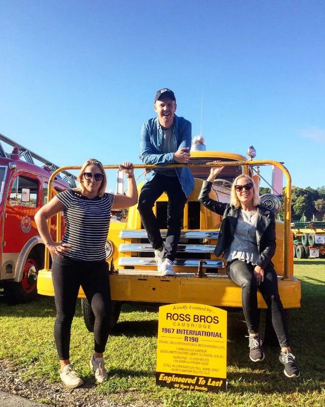 Three people posing with a vintage yellow fire truck labeled "Ross Bros Cambridge 1967 International R190".