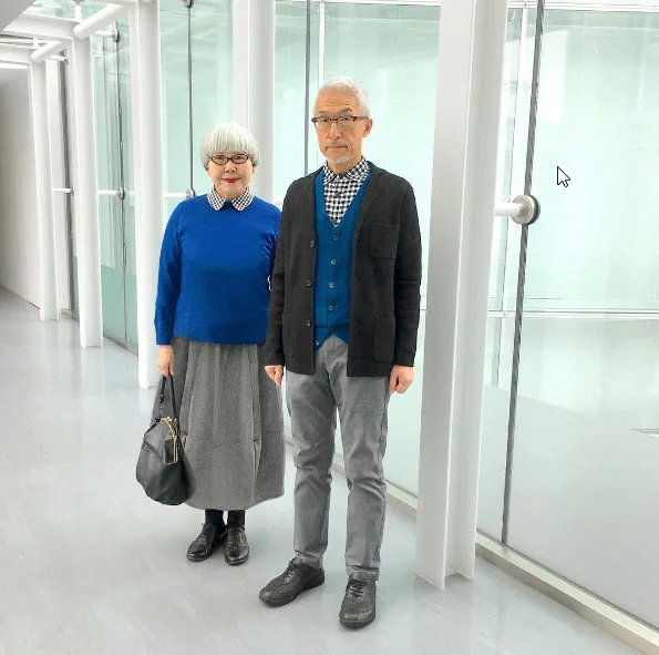Elderly couple in matching blue and black outfits standing in a modern glass hallway.