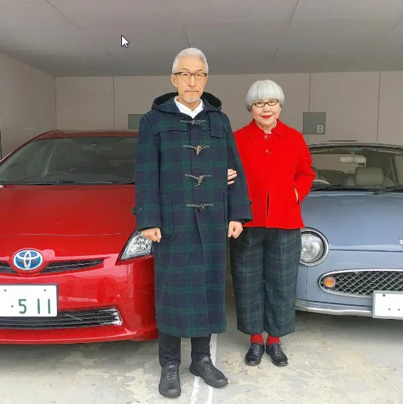 Elderly couple in stylish attire stands between red and blue cars in a parking area.