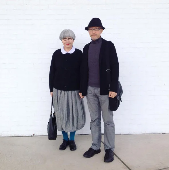 Elderly couple in coordinated black outfits stands against a white brick wall, holding hands and wearing glasses.