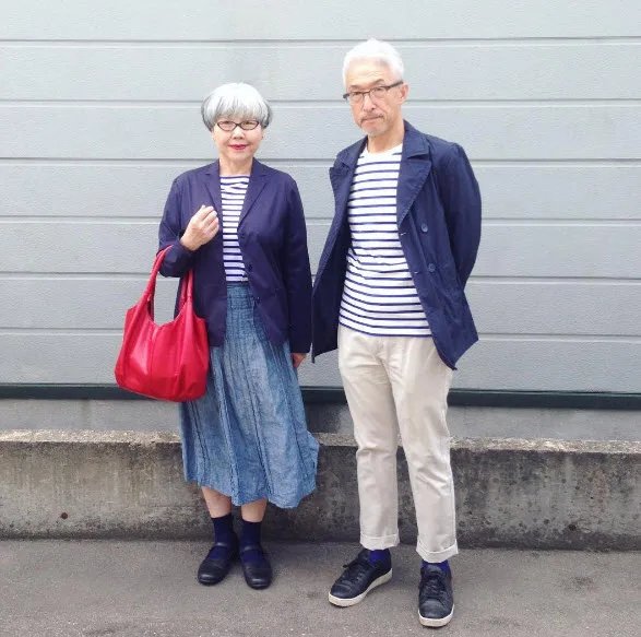 Elderly couple in matching navy and striped outfits, woman holding a red bag, standing against a light gray wall.
