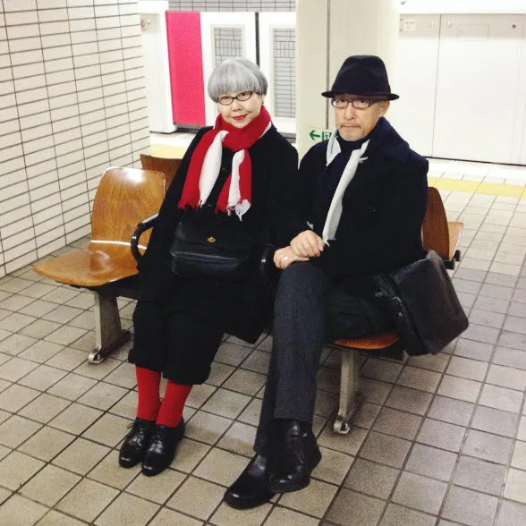 Elderly couple in stylish black coats and red scarves sitting on subway bench.