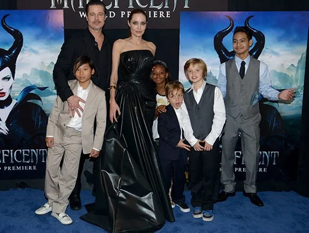A group of people, including two adults and several children, pose on a blue carpet at a "Maleficent" premiere event.