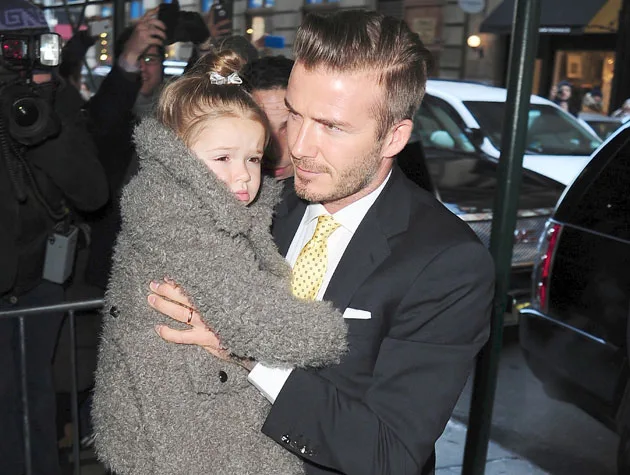 Man in suit holding a child in a fuzzy coat while facing photographers outside a building.