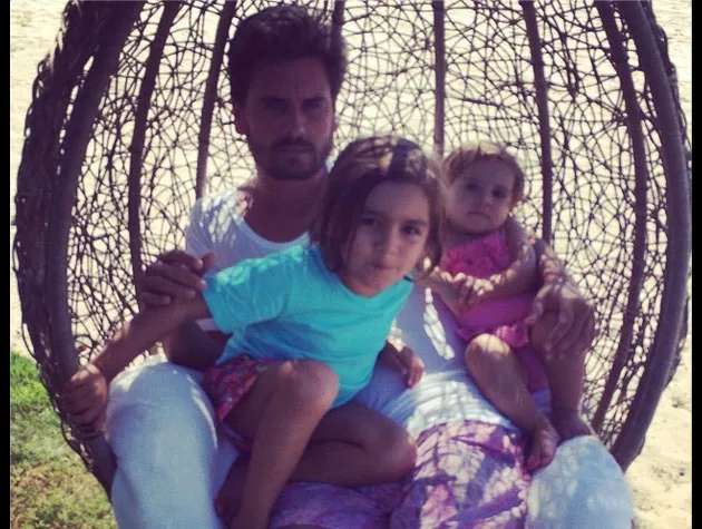 Man sitting in a wicker swing chair on a beach with two young children, one wearing a blue shirt and the other in pink.