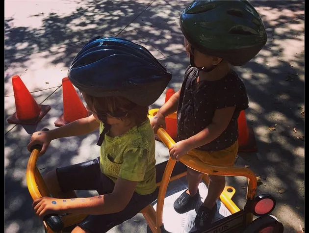 Children wearing helmets ride a small bike taxi, surrounded by orange traffic cones on a sunny day.