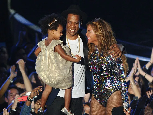 A family embracing on stage, woman in sequined outfit, man holding a child in a gold dress, audience applauding.
