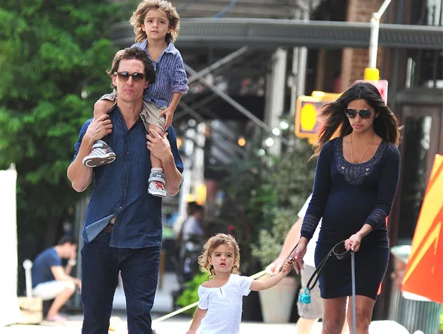 A family walking in the city; man carries a child on shoulders, woman holds hands with another child, urban setting.