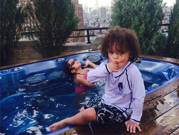 Two children in swimwear by a rooftop hot tub, with city buildings in the background.