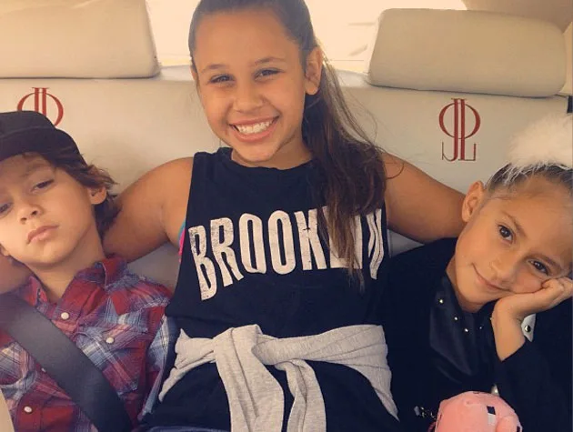 Three children sitting in a car. The center child is smiling broadly, wearing a "Brooklyn" shirt.