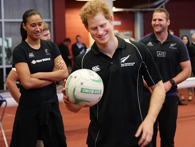Man with red hair holding a rugby ball, smiling, surrounded by others in a sports facility.
