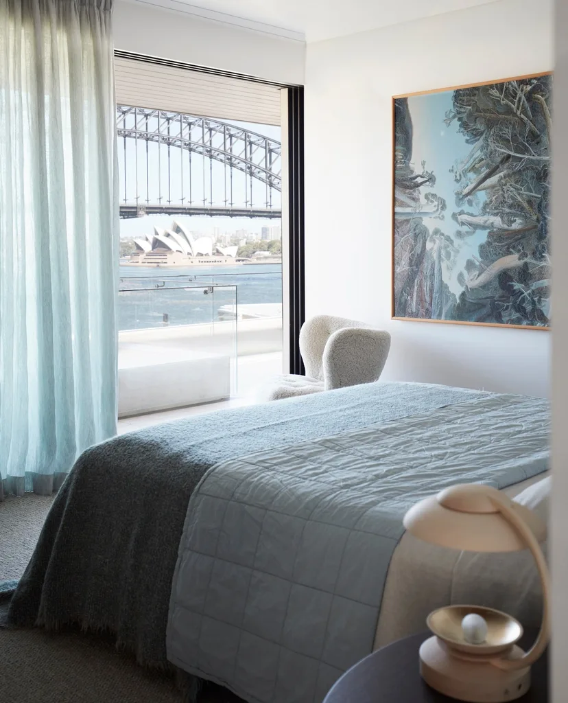 Pale blue sheer curtains frame a bedroom window looking out towards the Sydney Harbour Bridge and Opera House.