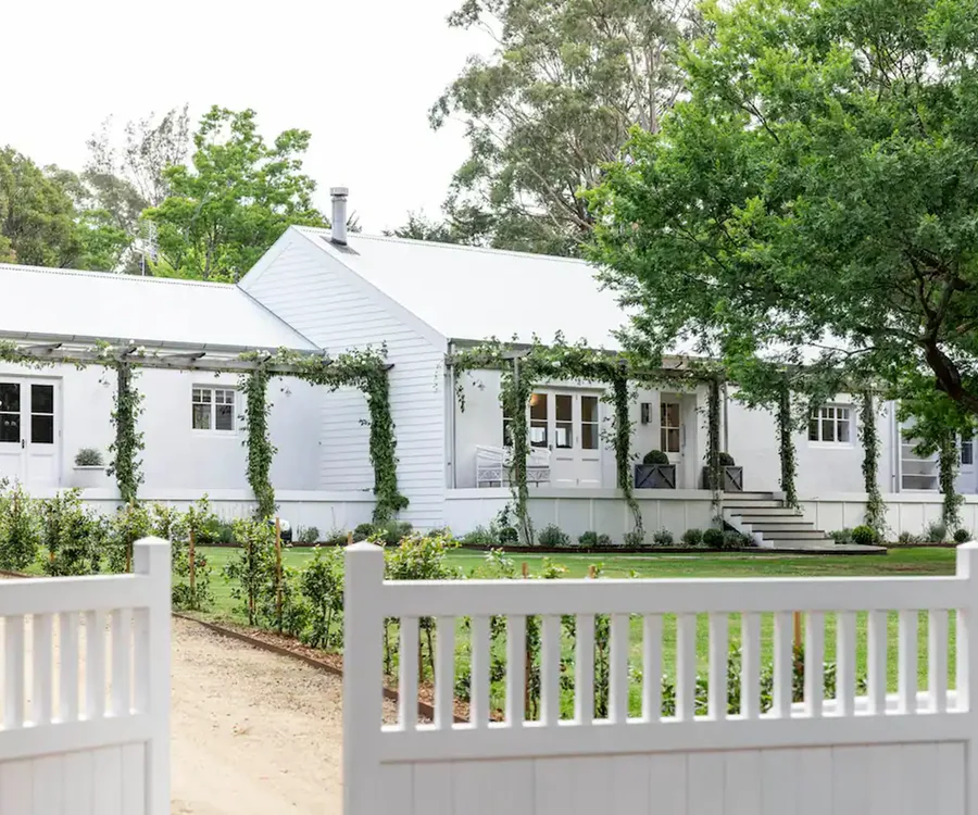 White house with greenery and vines, set behind a white picket fence, surrounded by lush trees and garden.
