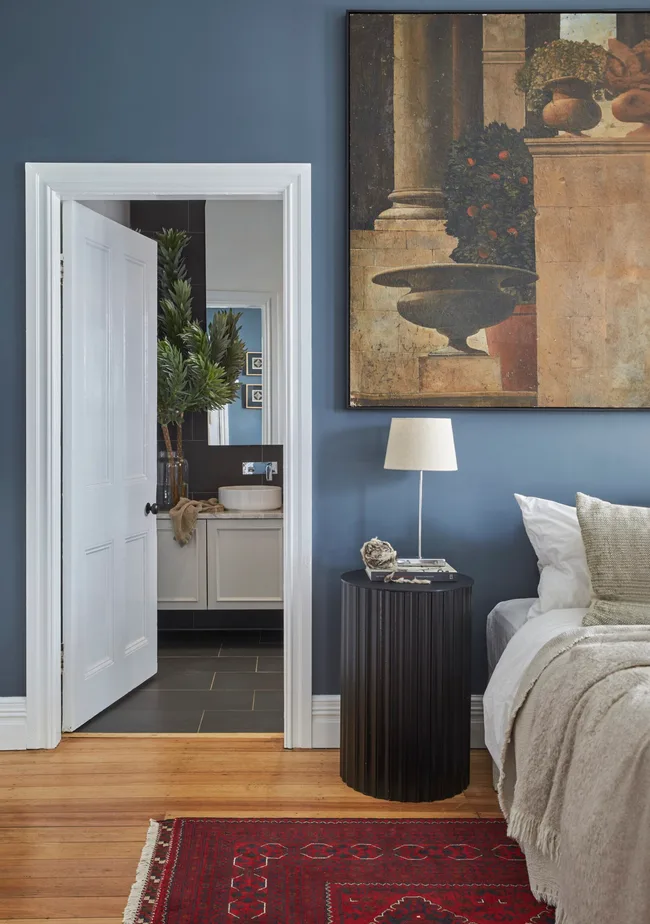 A blue bedroom with a large painting hanging over a bed with neutral-toned bedding