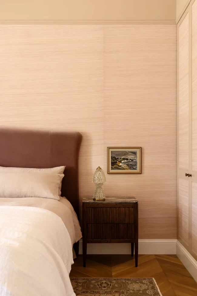 A bedroom with pale pink wallpaper, a velvet bedhead and timber bedside table