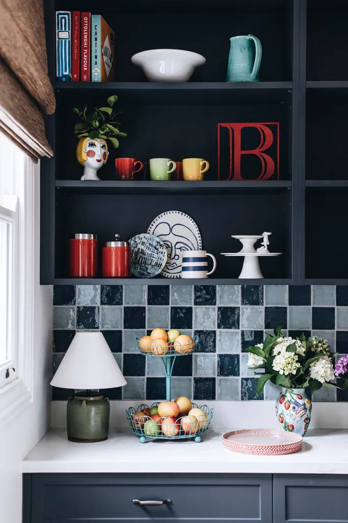 A kitchen storage space filled with mugs and fruit, with a blue checkerboard splashback