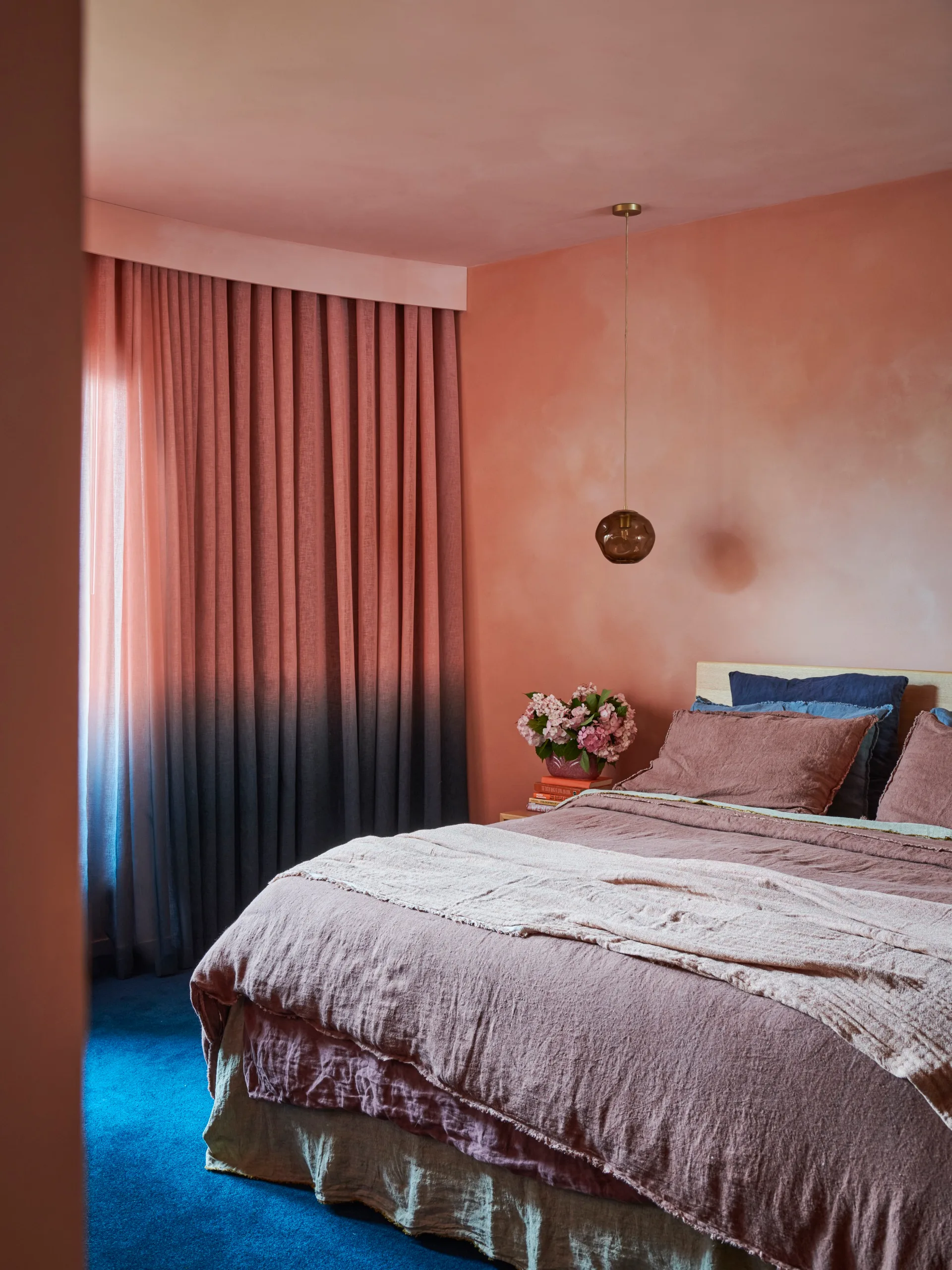 A bedroom with coral coloured limewash walls, ombre curtains that go from blush to blue, blending into the blue carpet.