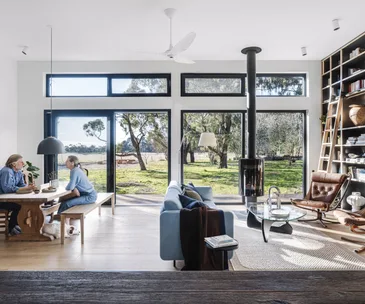 A couple sit at a wooden dining table with a dog at their feet. The room also contains a blue sofa, brown leather chairs, a contemporary fireplace and a large, floor-to-ceiling library. Large windows open the space onto a grassy rural outlook.