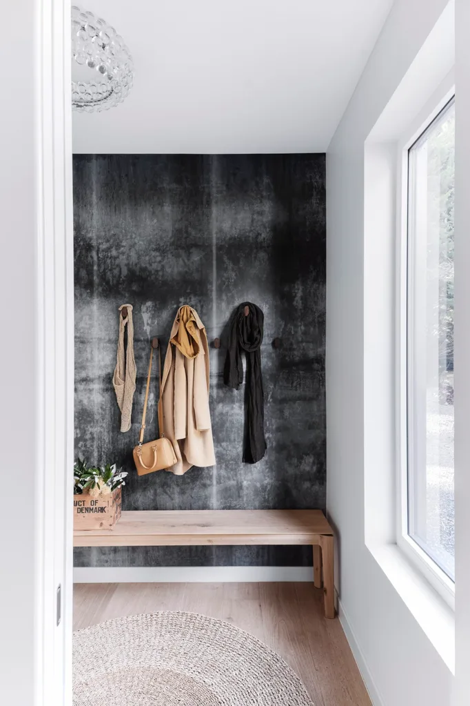 An entryway with a textured black wall and coats hanging from wooden pegs. A jute rug sits atop floorboards.