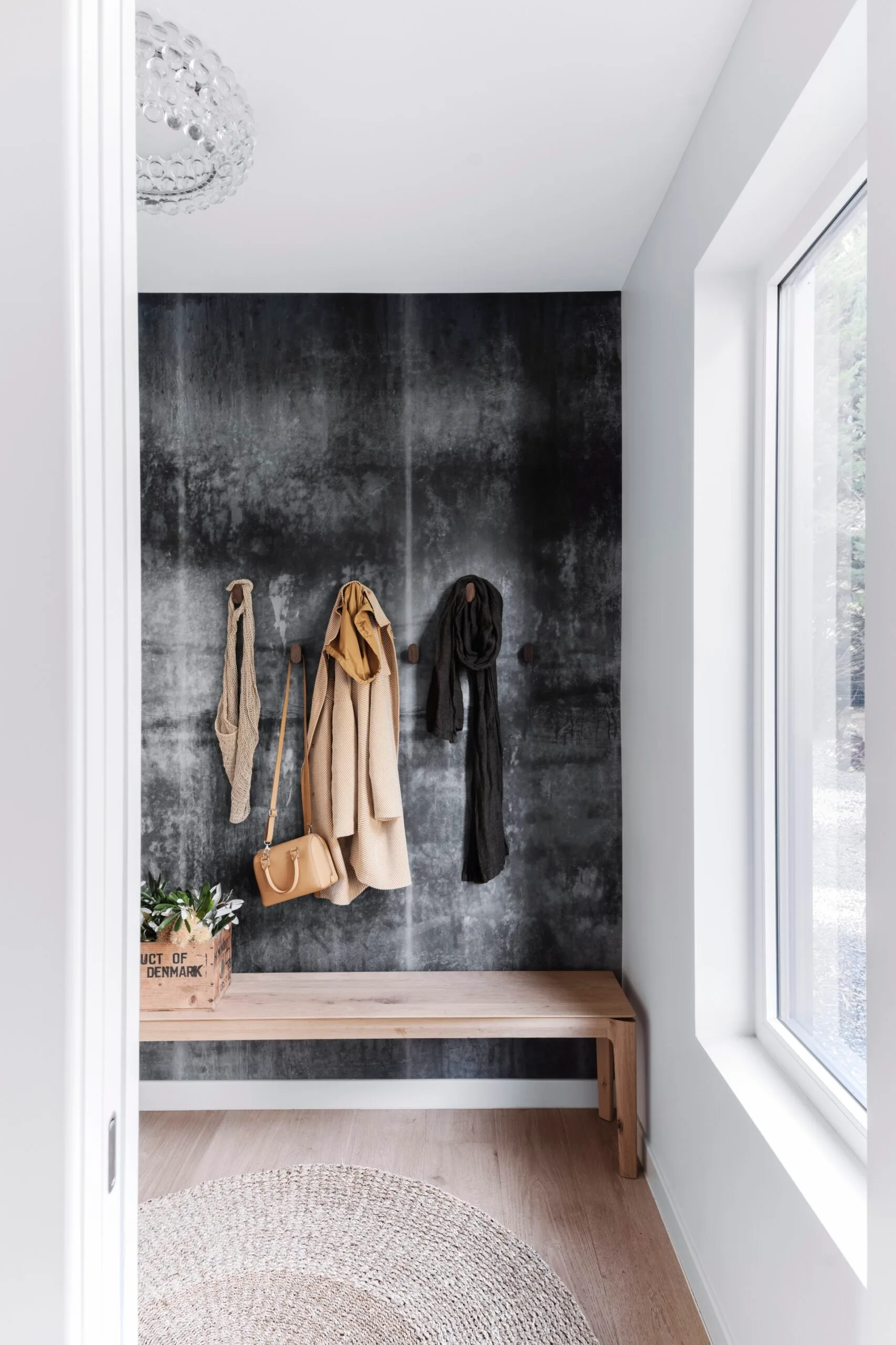 An entryway with a textured black wall and coats hanging from wooden pegs. A jute rug sits atop floorboards.