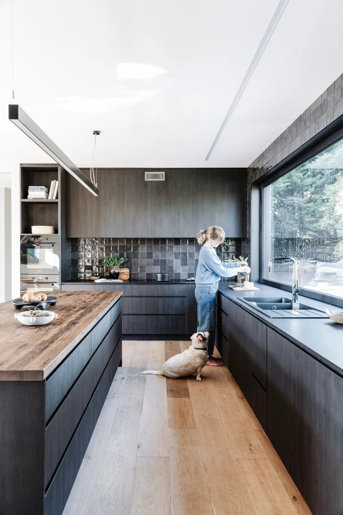 A women stands in a black kitchen with glossy tiles, wooden floorboards and a wooden island benchtop.