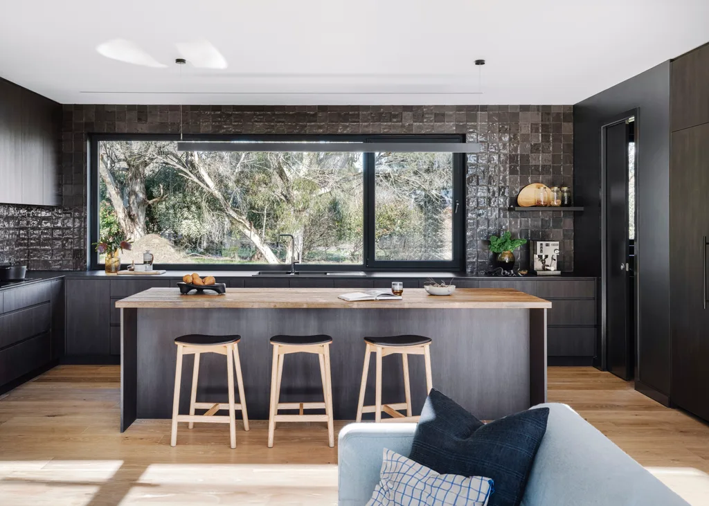 A black kitchen with glossy, square tiles, a wooden island bench and wooden stools.