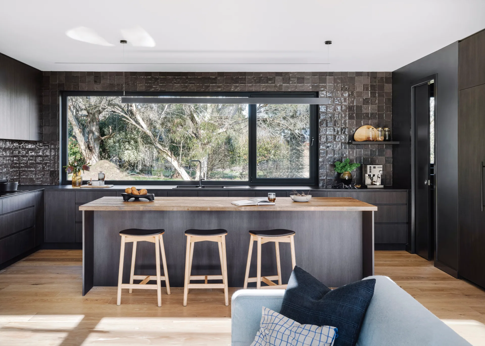 A black kitchen with glossy, square tiles, a wooden island bench and wooden stools.