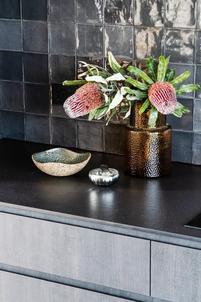 A small vignette of banksia flowers in an amber vase, a ceramic bowl and a salt dish in a black kitchen.
