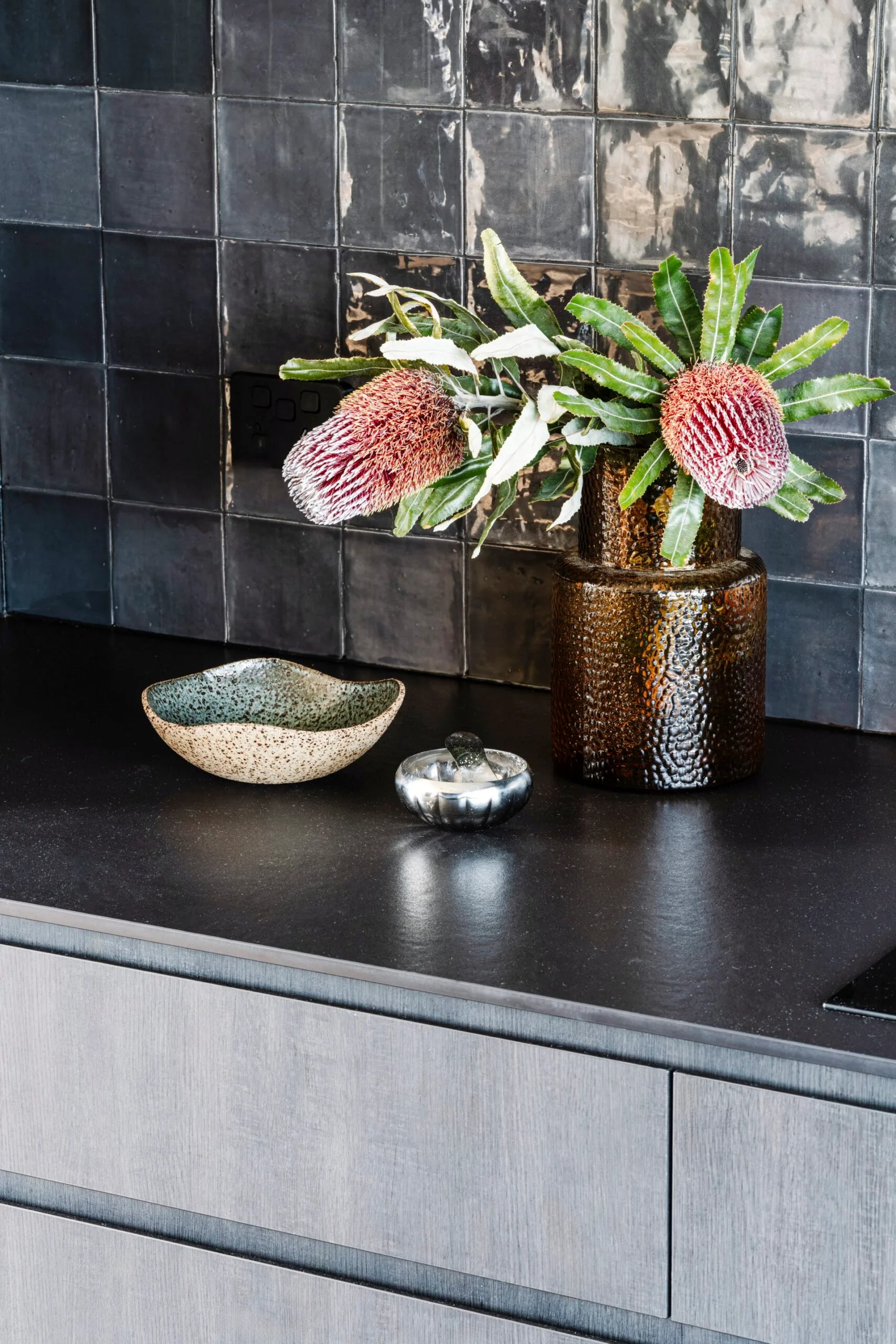 A small vignette of banksia flowers in an amber vase, a ceramic bowl and a salt dish in a black kitchen.