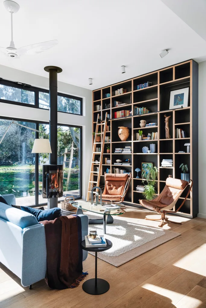 A large, floor-to-ceiling black bookcase sits along one wall of a living room. A large glass window, contemporary fireplace and brown leather armchairs complete the space.