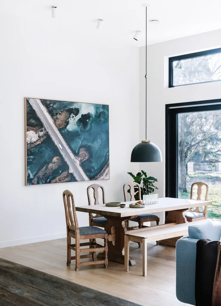 A wooden dining table with a black pendant light hanging overhead. On the wall behind there is a coastal photograph.