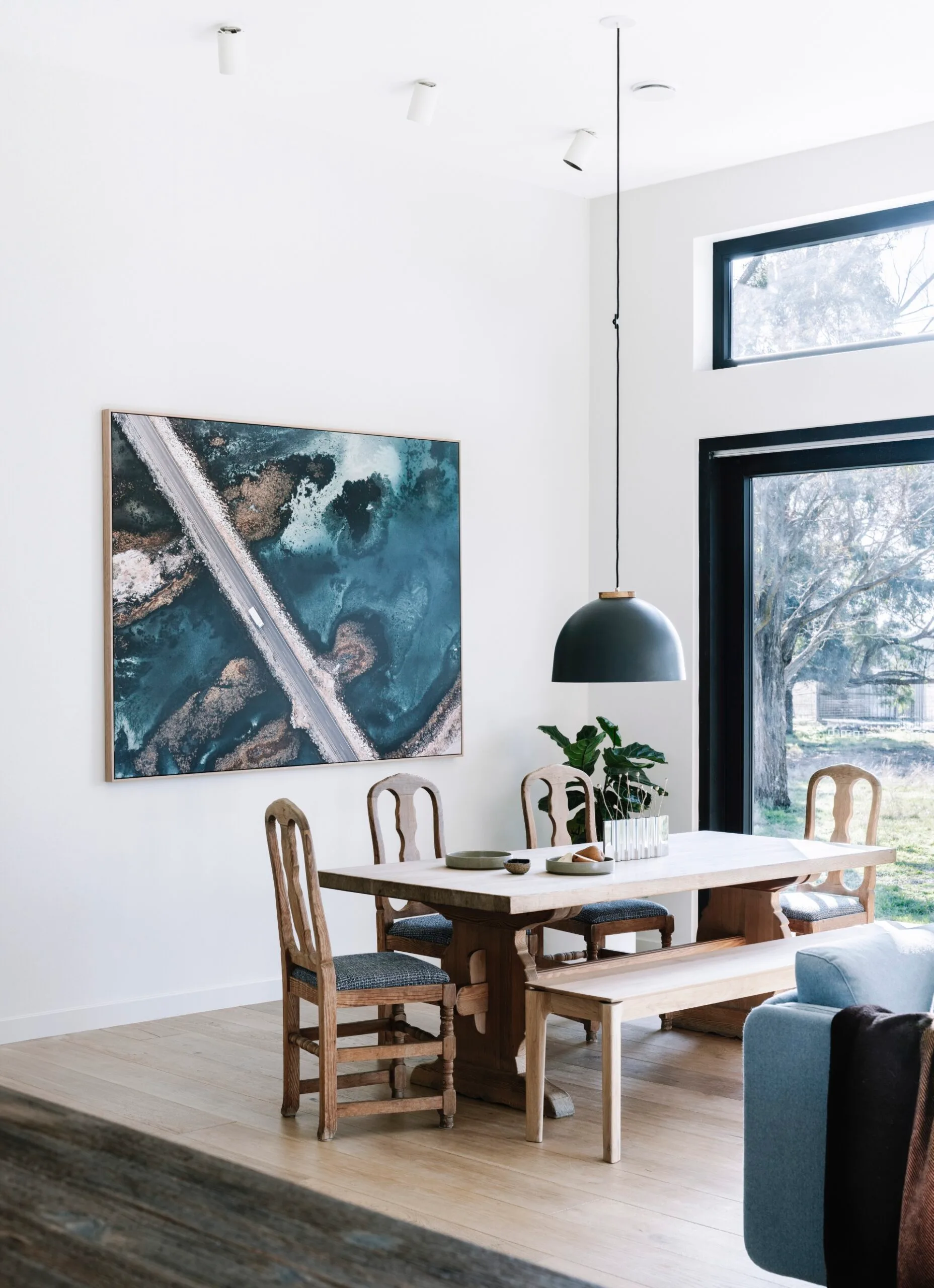 A wooden dining table with a black pendant light hanging overhead. On the wall behind there is a coastal photograph.