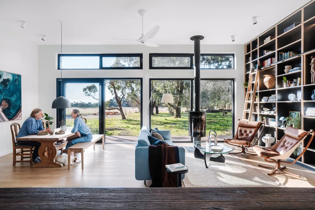 A couple sit at a wooden dining table with a dog at their feet. The room also contains a blue sofa, brown leather chairs, a contemporary fireplace and a large, floor-to-ceiling library. Large windows open the space onto a grassy rural outlook.