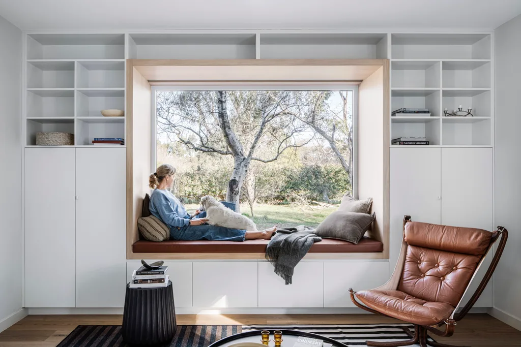 A woman sits on a window seat looking out onto Australian bushland. In the foreground, a lounge room is styled with a brown leather chair and striped rug.