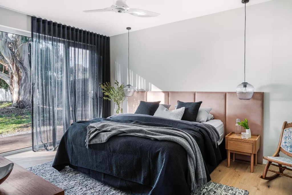 A bedroom with black sheer curtains, black bedding and a brown, upholstered bedhead. Glass pendant lights hang over wooden bedside tables, while a black and white rug covers a large section of the wooden floor.