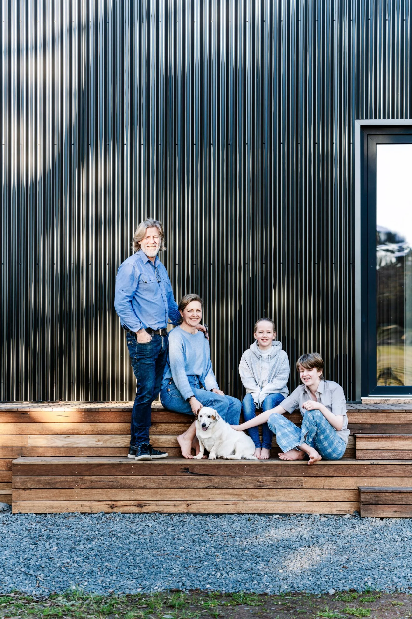 A family and their white dog sit on wooden steps in front of a black, corrugated-iron-clad home designed with Passivehaus principles.
