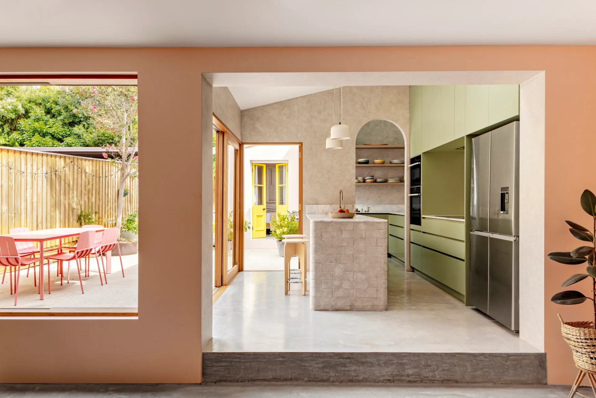 A kitchen with green cabinets looks out to a sunny alfresco dining area with a pink outdoor dining set