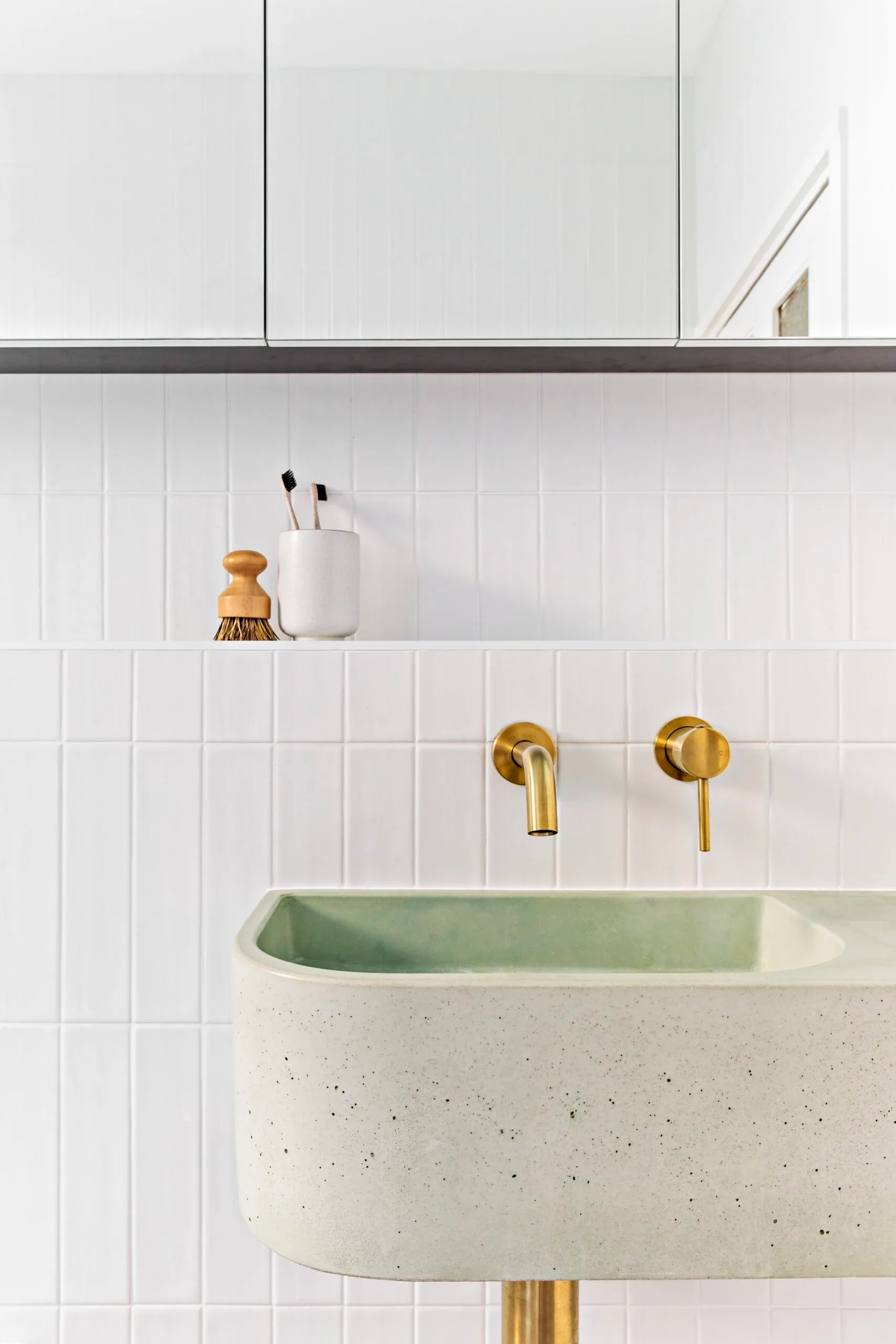 A white tiled bathroom with a subtly green concrete sink and gold fittings