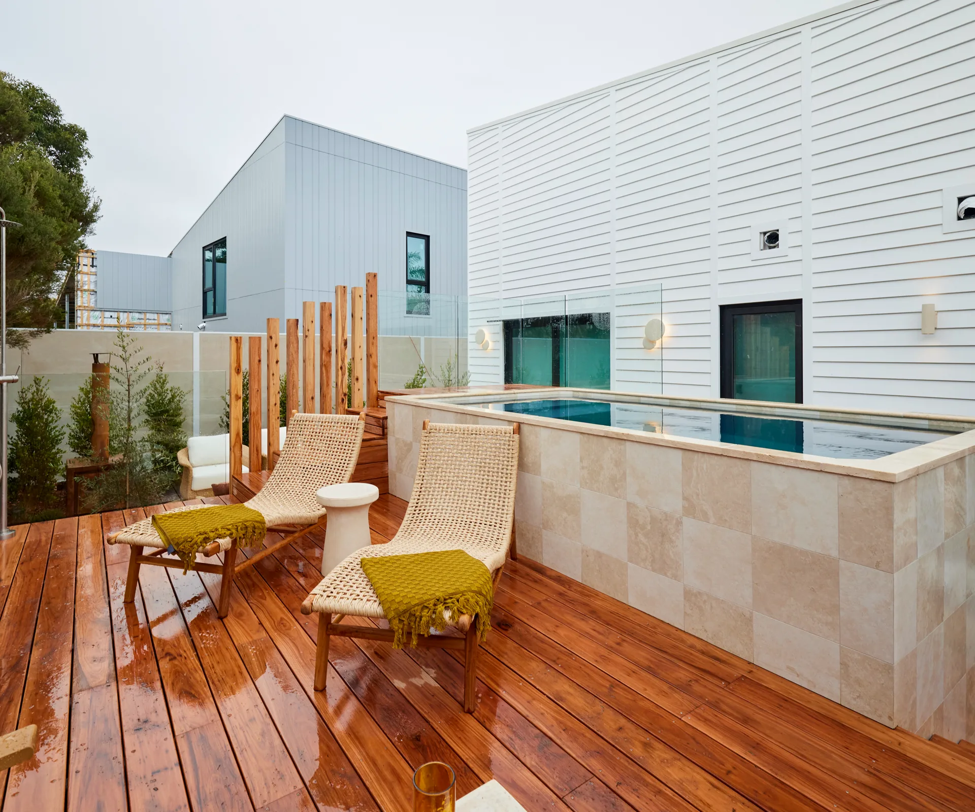 An above-ground pool with a checkerboard tile pattern, two rattan daybeds and an outdoor shower.