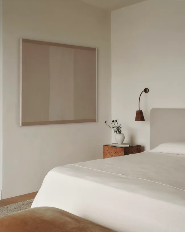 Neutral bedroom with white bedding, a velvet foot stool and rattan wall sconce