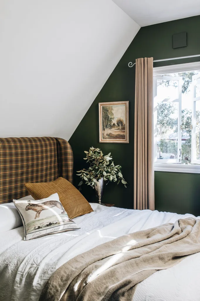 A bedroom with a slanted ceiling, a dark green wall and a mustard, tartan upholstered headboard. The bed is made with white bedding with brown cushions and a throw.