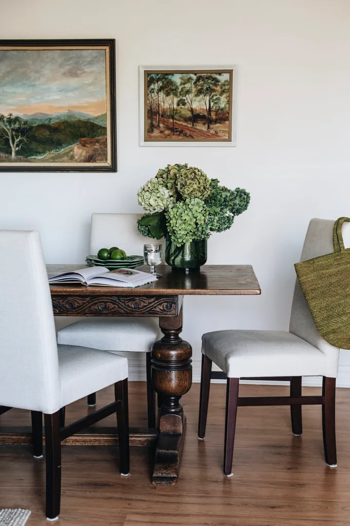 An antique wooden dining table is surrounded by white upholstered chairs and topped with a vase of green hydrangeas.
