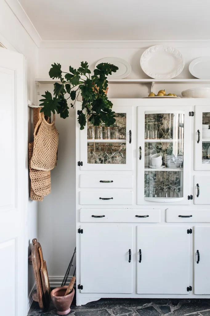 A white, antique crockery cabinet with black handles. It is filled with glassware and crockery.